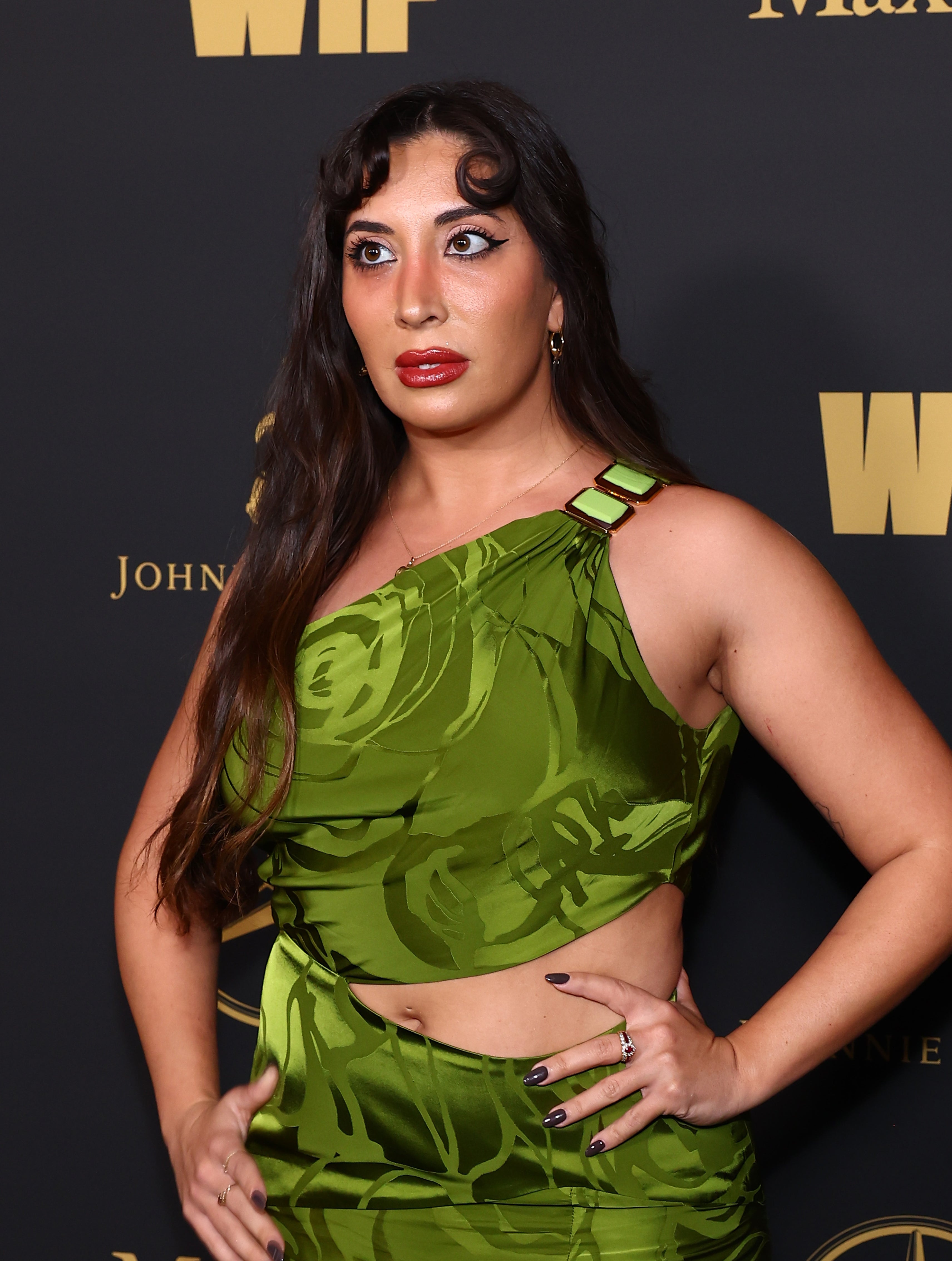 Sarah posing in a patterned front-cutout dress with hands on hips against a logo backdrop at a media event