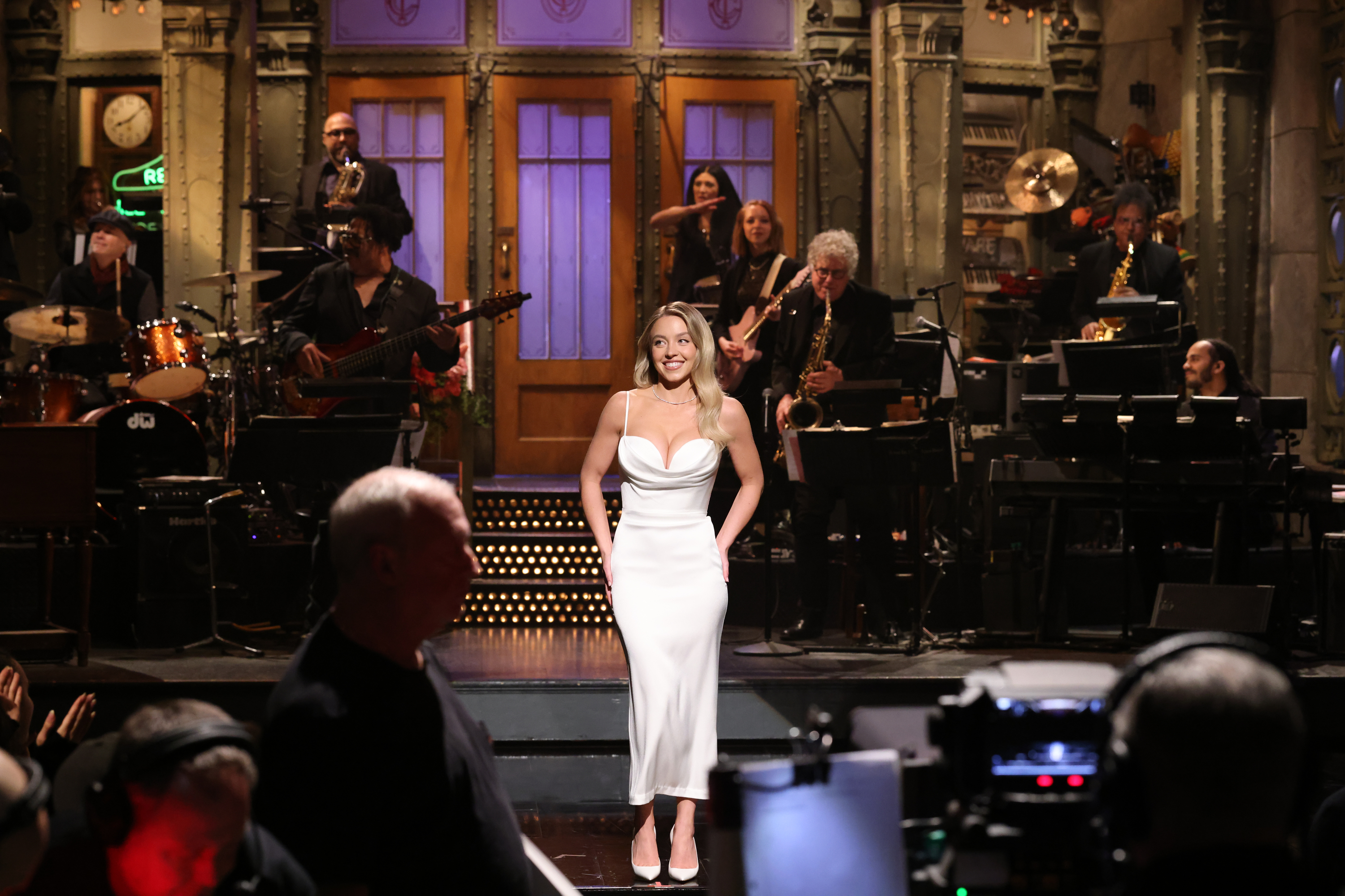 Sydney in a spaghetti-strap dress on the SNL stage with band in background