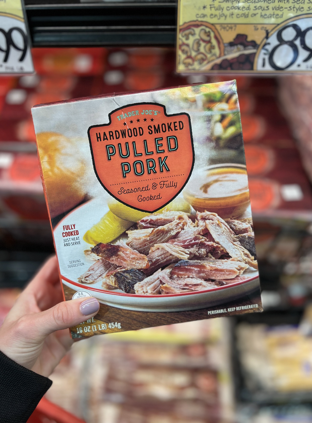 Hand holding a package of Trader Joe&#x27;s Hardwood Smoked Pulled Pork in a grocery store aisle