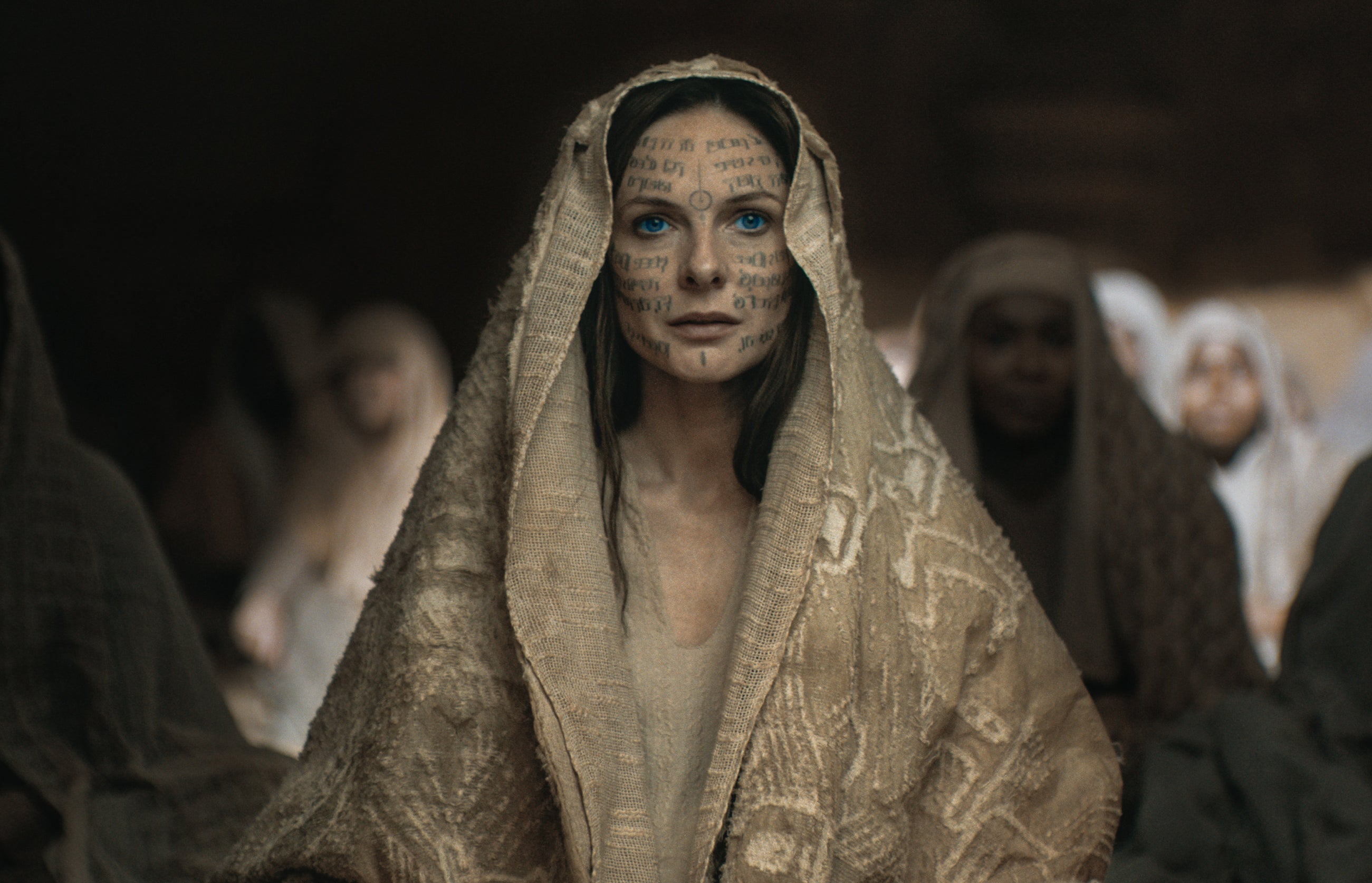 Rebecca Ferguson in character as Jessica with ceremonial facial markings, draped in a textured shawl, with extras in the background