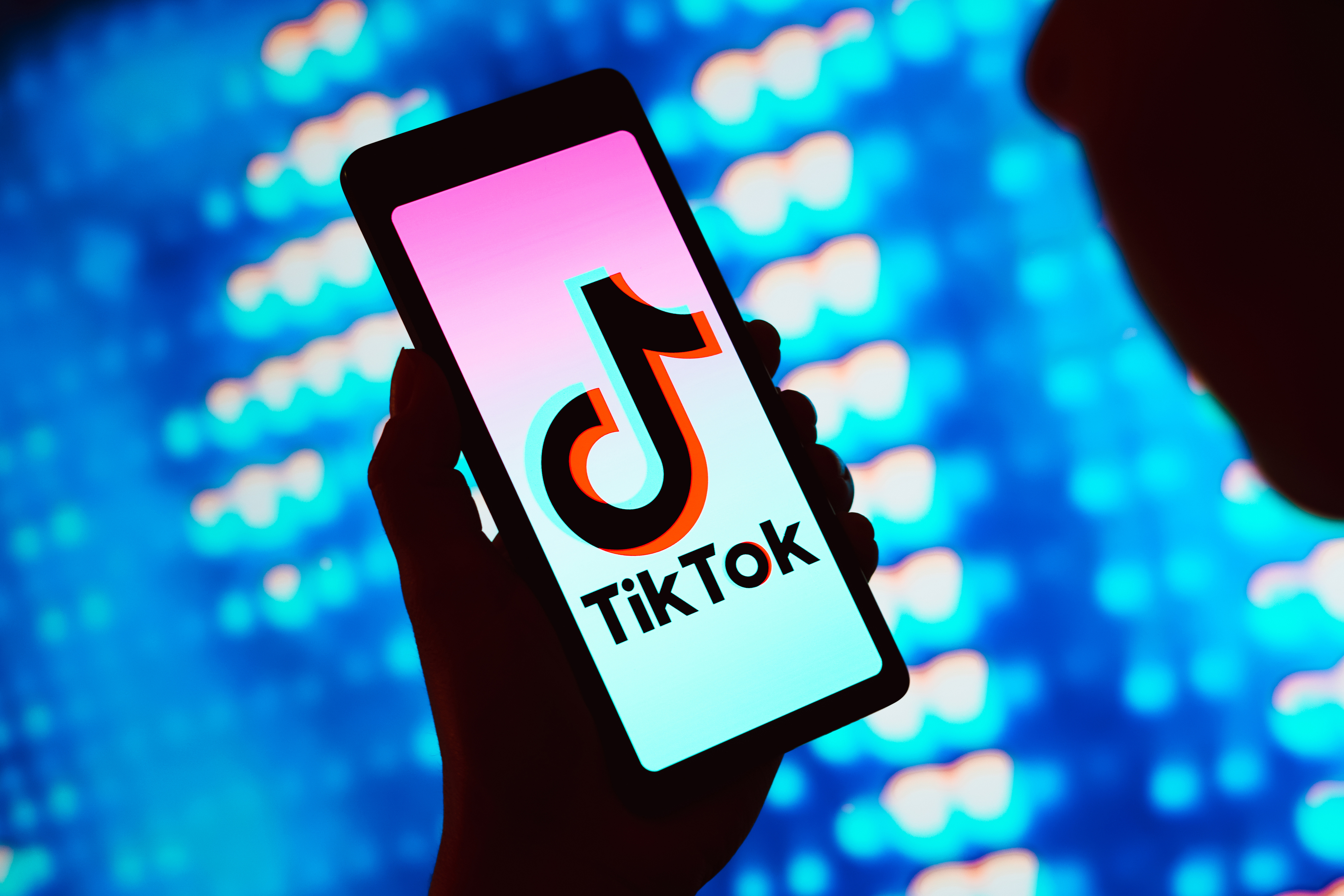 Silhouetted person holding a smartphone with the TikTok app logo on the screen, illuminated background