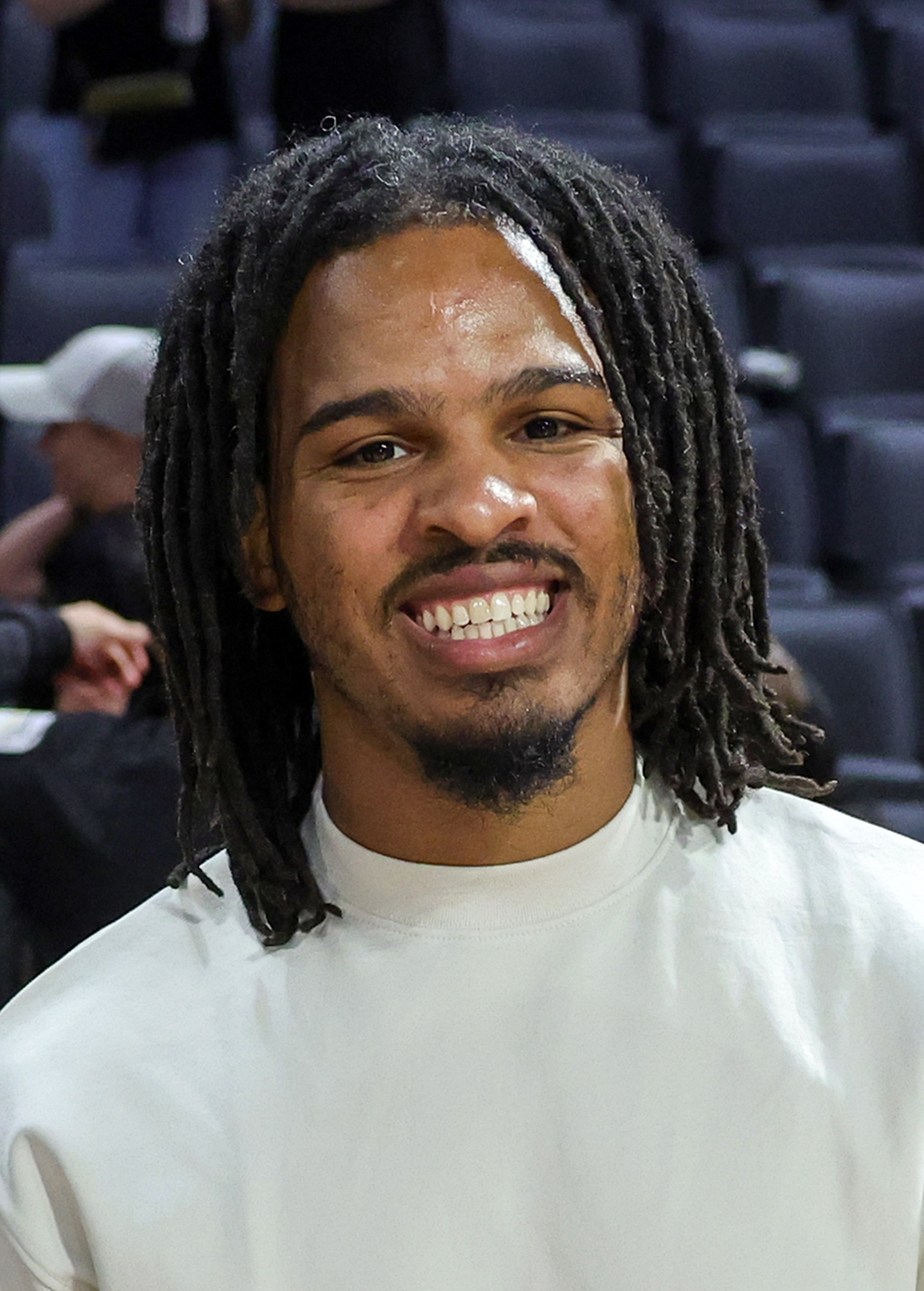 Man with dreadlocks smiling at the camera, wearing a plain shirt, at an indoor event