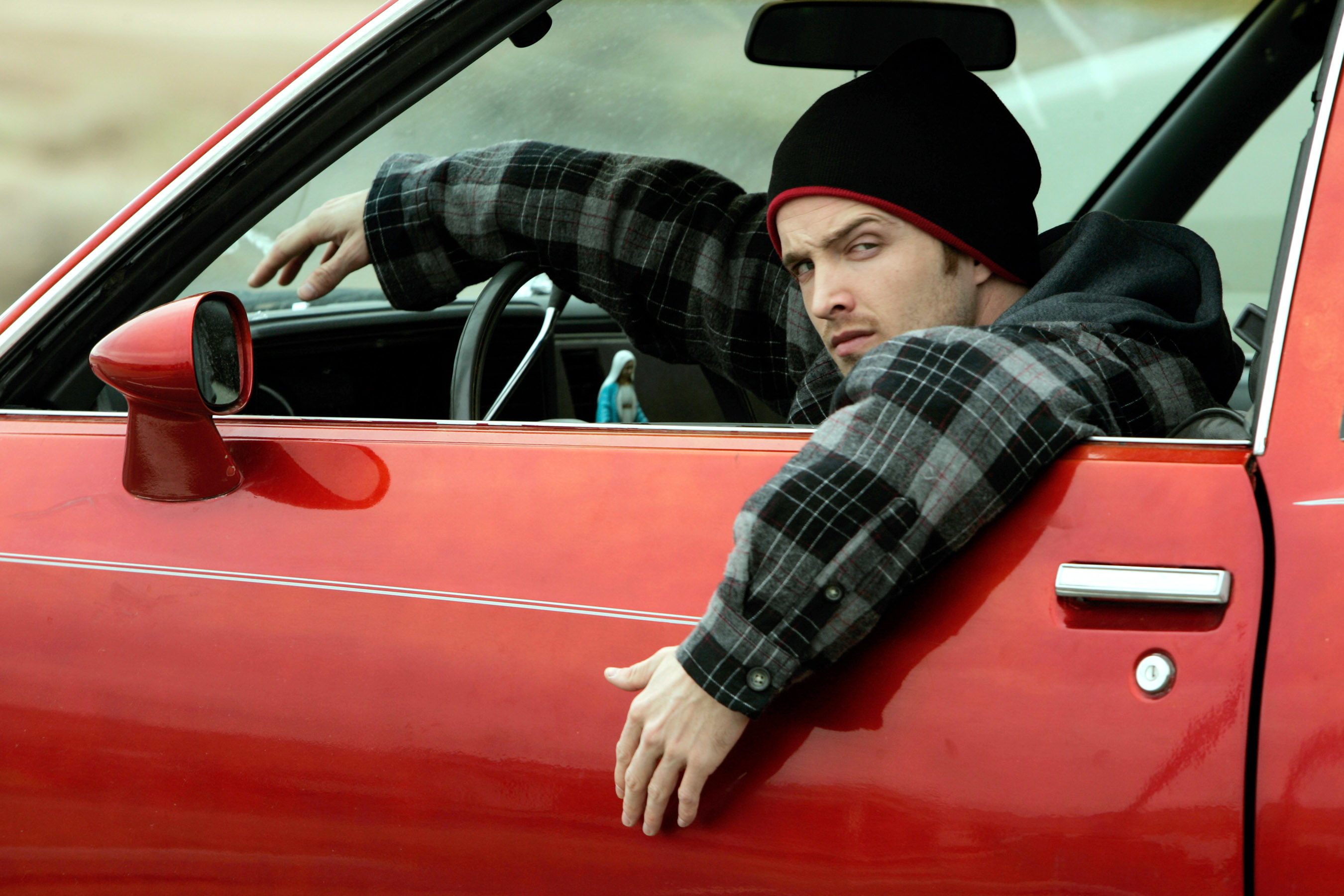 Jesse Pinkman, wearing a beanie and plaid jacket, looks concerned while leaning out of a red car window