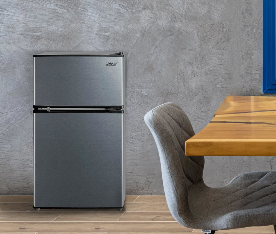 Gray fridge next to a wooden table and a gray chair in a room with a concrete wall