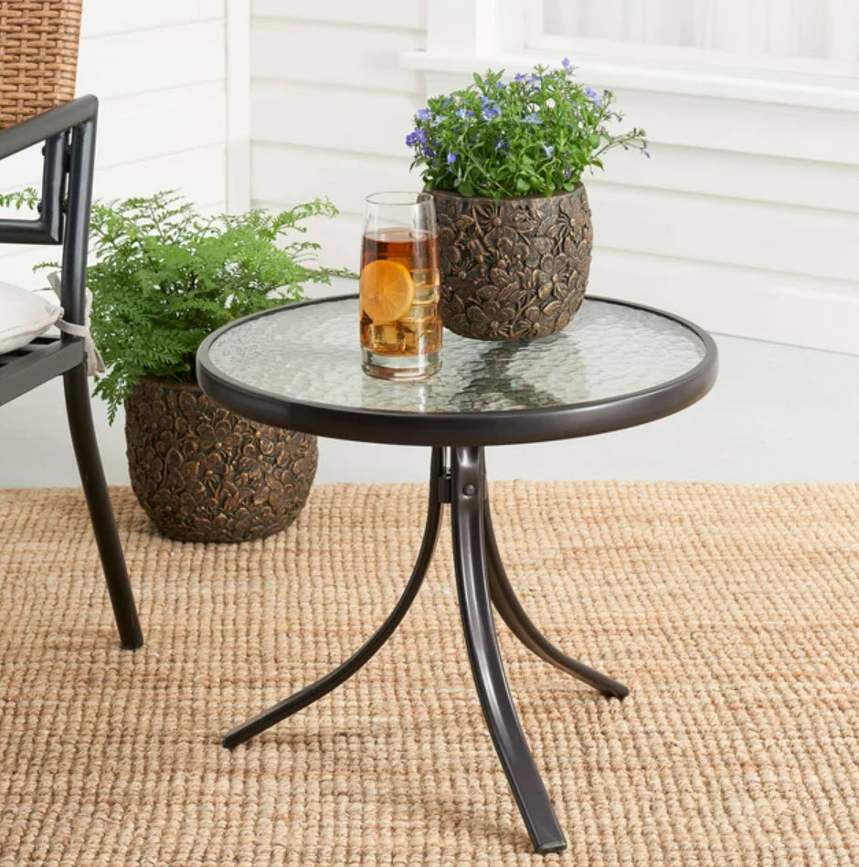A round outdoor table with a glass on top and two decorative potted plants, placed on a textured rug