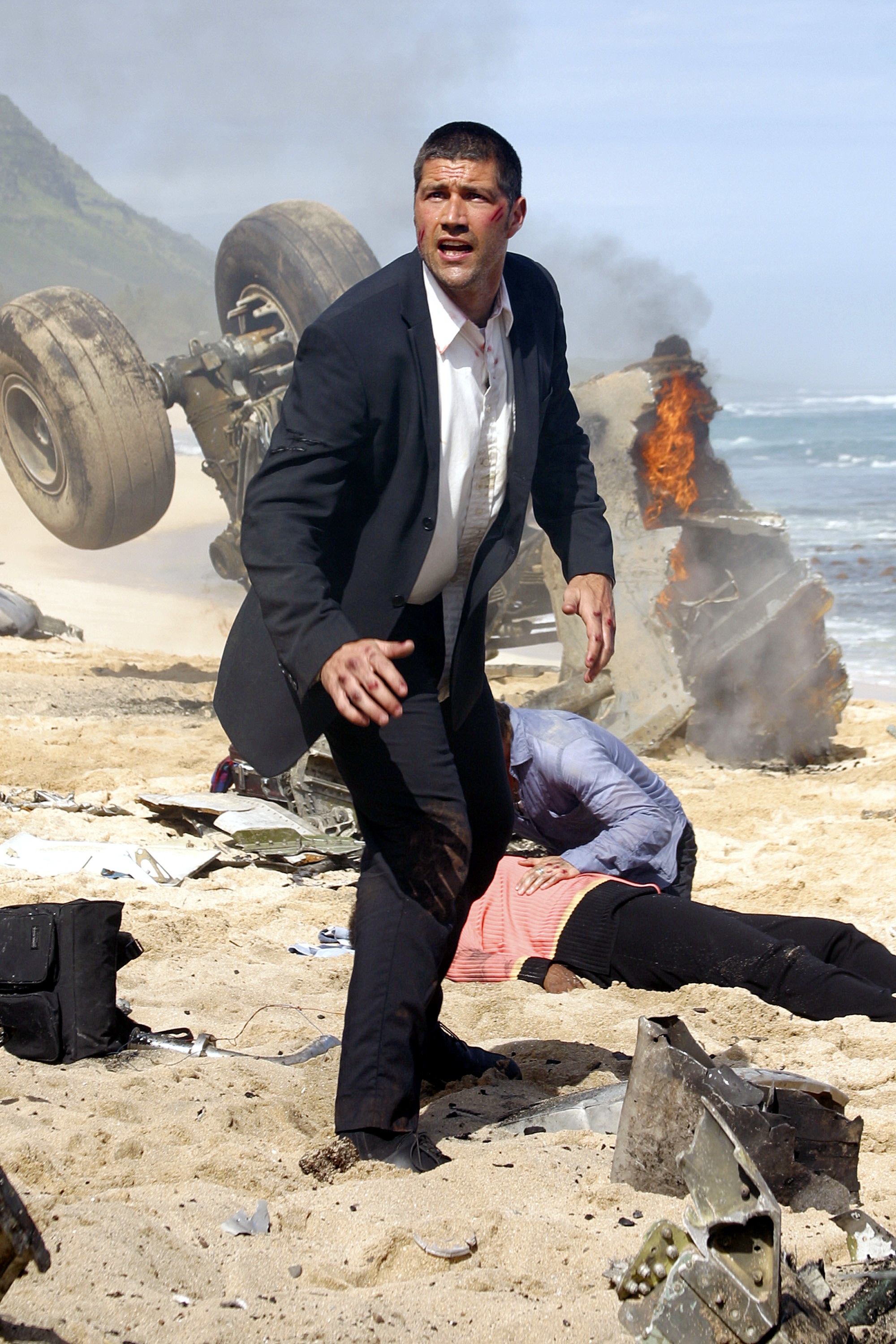 A man in a suit running from a fiery crash site with a large tire and debris around