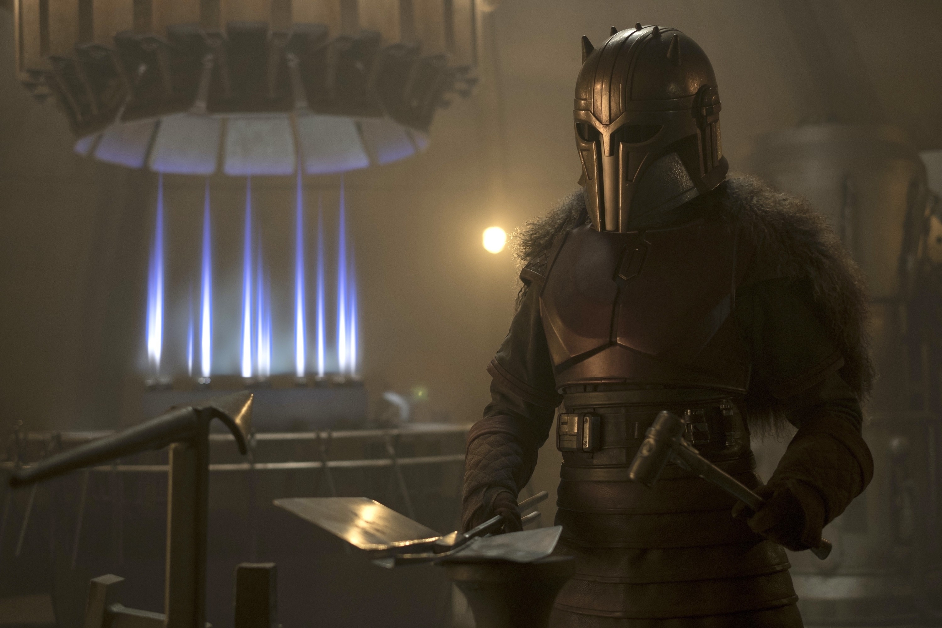 The Mandalorian in armor stands beside a forging table, holding a piece of beskar