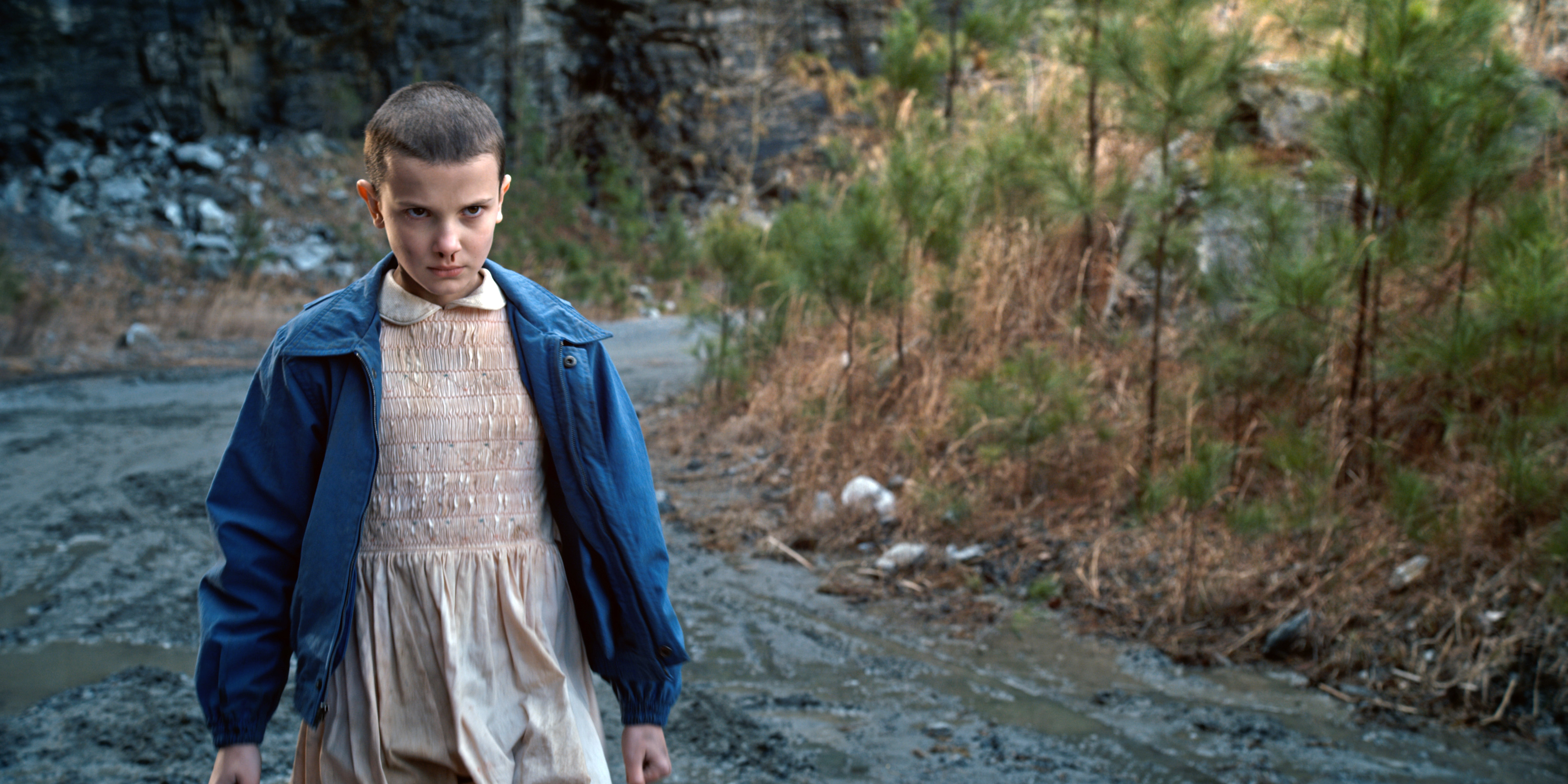 Eleven from Stranger Things walking outdoors in a blue jacket and dress