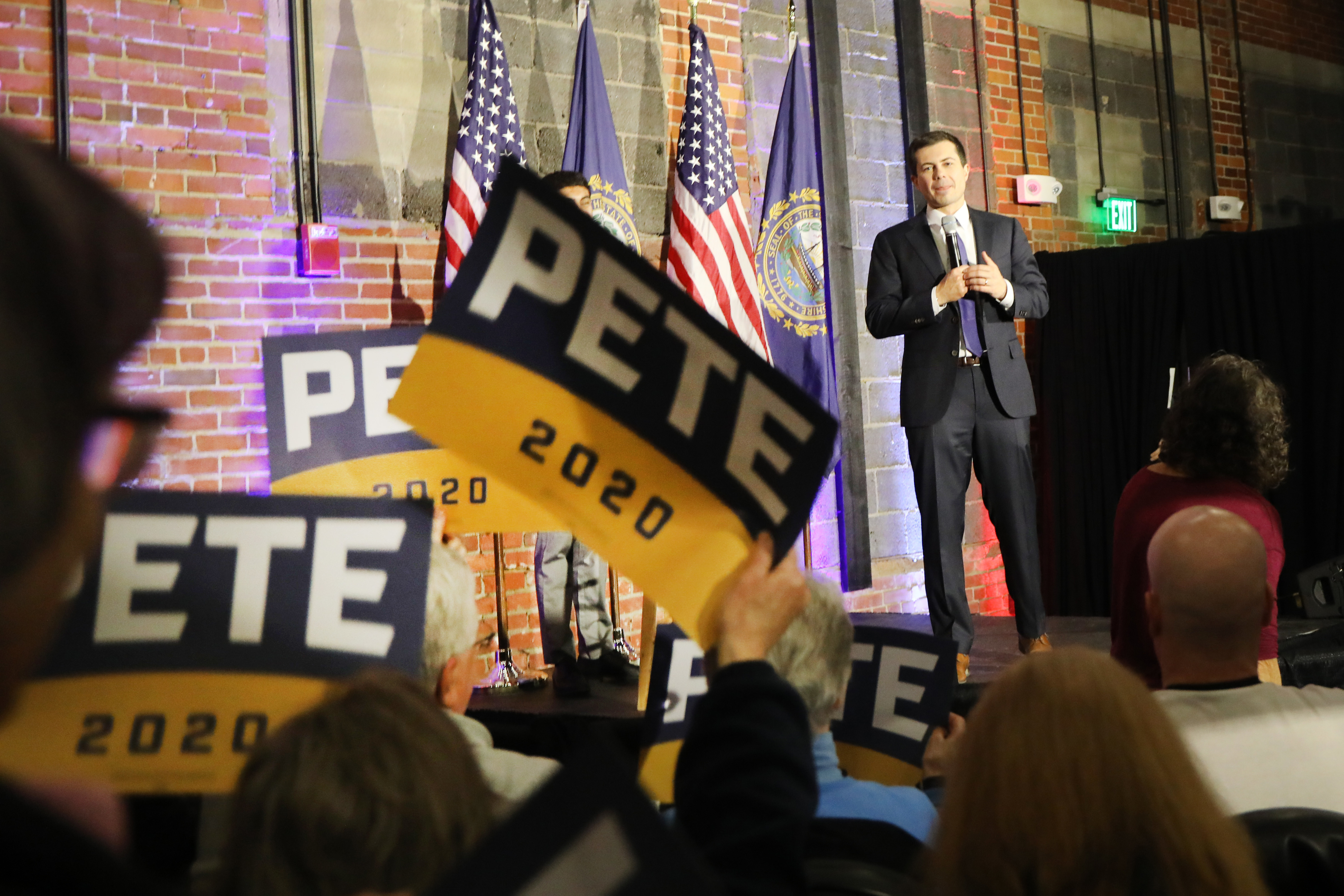 Pete Buttigieg stands speaking at a rally with supporters holding campaign signs