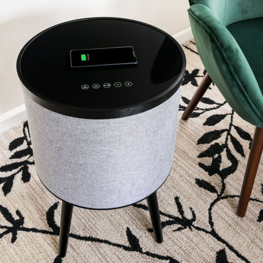 Smart speaker with digital display and fabric cover, placed on a carpet next to a green chair