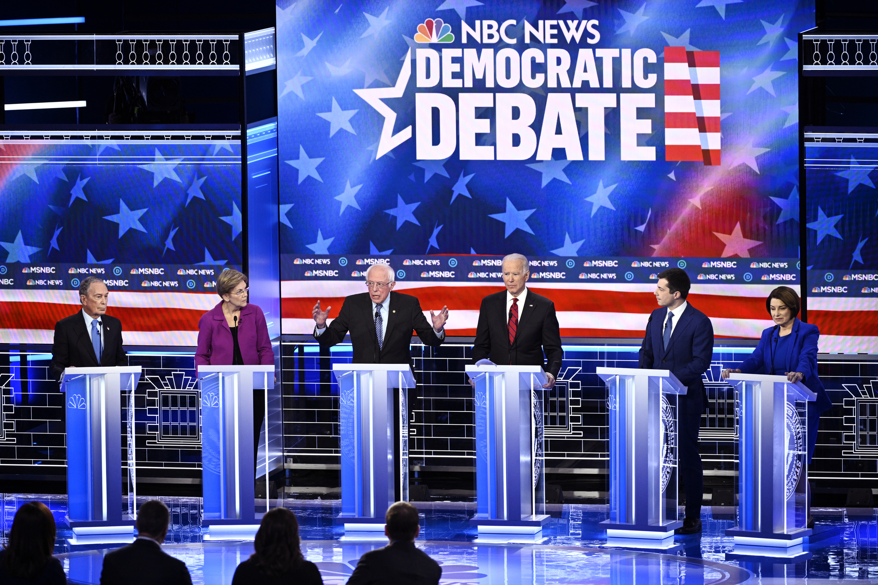 Six candidates — Mike Bloomberg, Elizabeth Warren, Bernie Sanders, Joe Biden, Pete Buttigieg, and Amy Klobuchar — at a Democratic debate onstage behind podiums with an NBC News backdrop