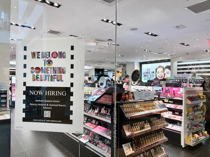 Storefront window with &quot;We Belong to Something Beautiful&quot; slogan and &quot;Now Hiring&quot; sign. Inside view of cosmetic product displays