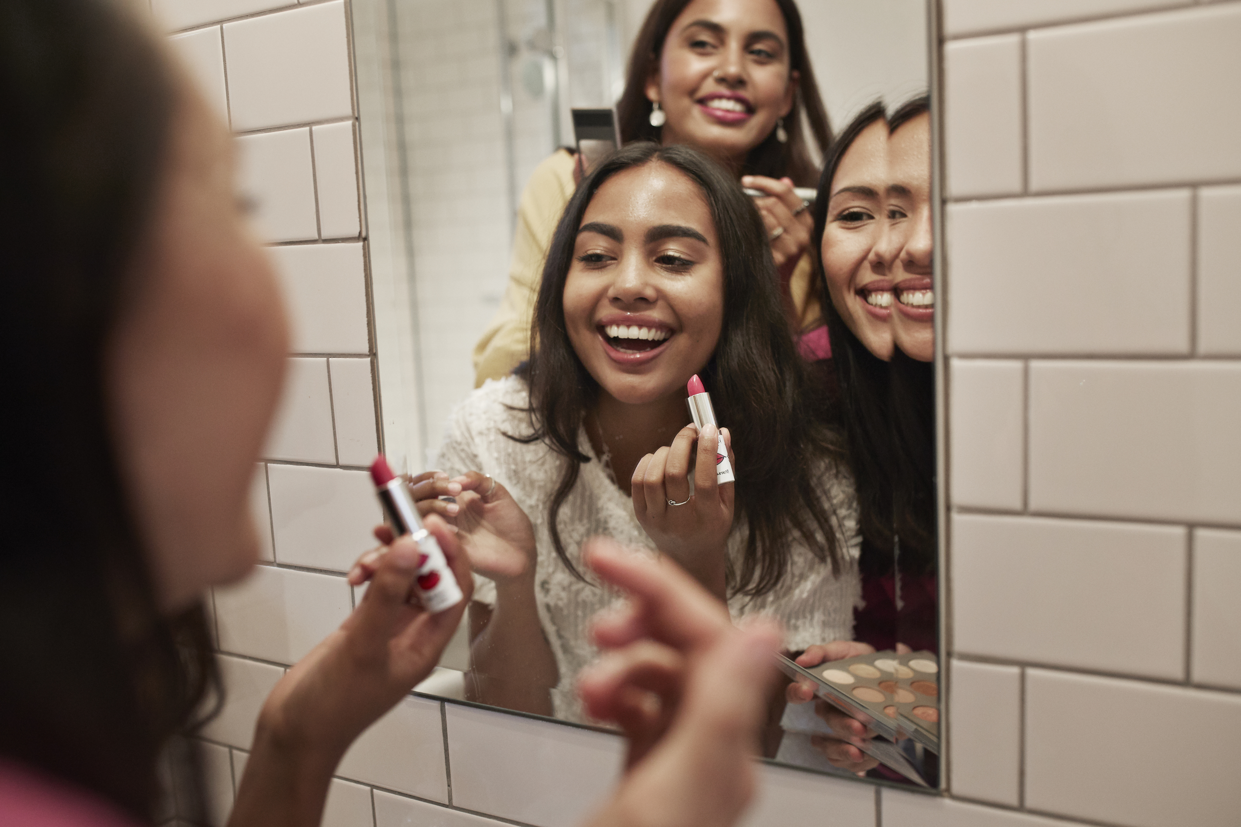 Three friends are smiling and applying makeup in a mirror