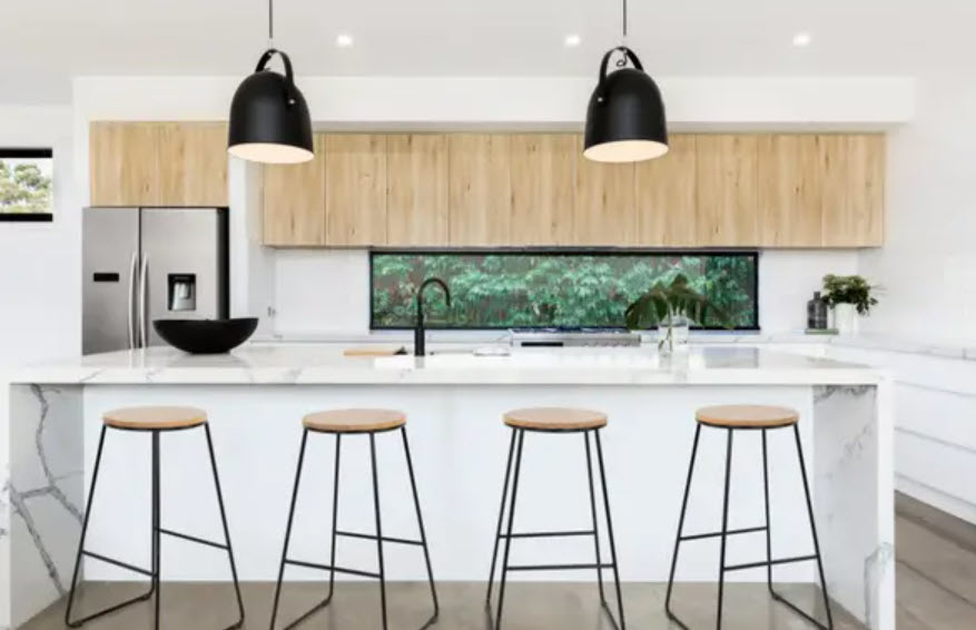 Modern kitchen interior with island, stools, and pendant lights, overlooking greenery through a horizontal window