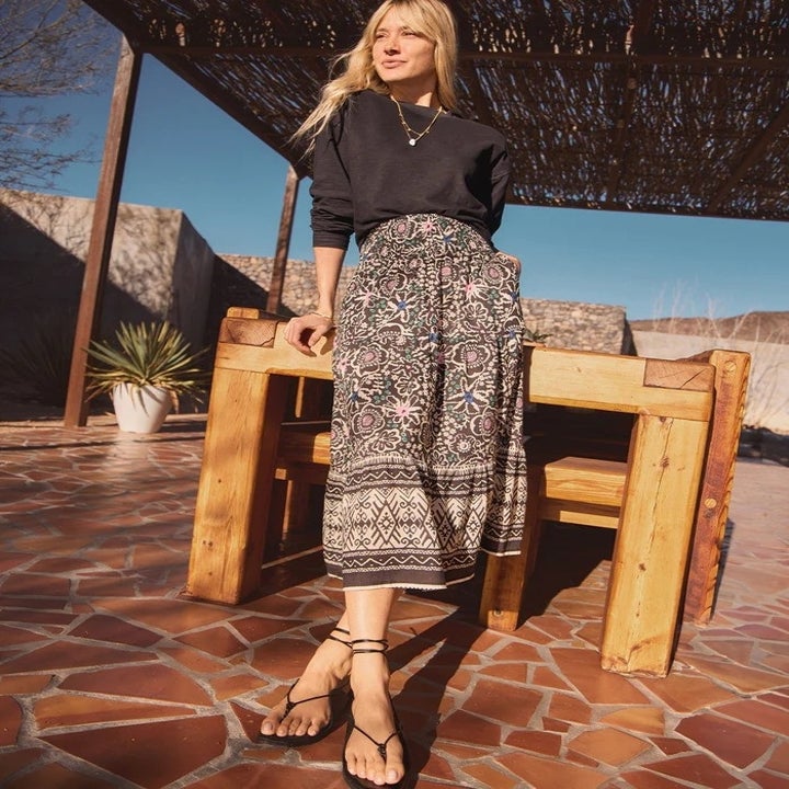 Woman in a bohemian-style skirt and black top posing outdoors near wooden chairs