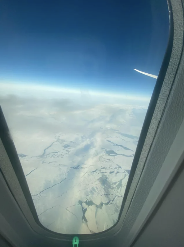 Aerial view of snowy landscape through airplane window, another plane&#x27;s contrail in the distance