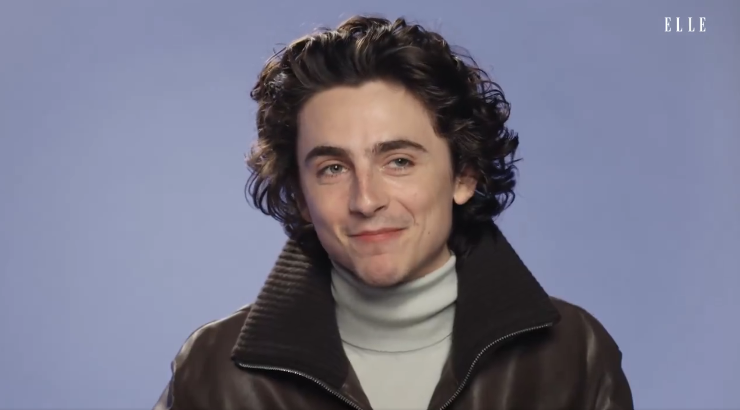 Timothée Chalamet with curly hair in a layered turtleneck and jacket, posing for the camera