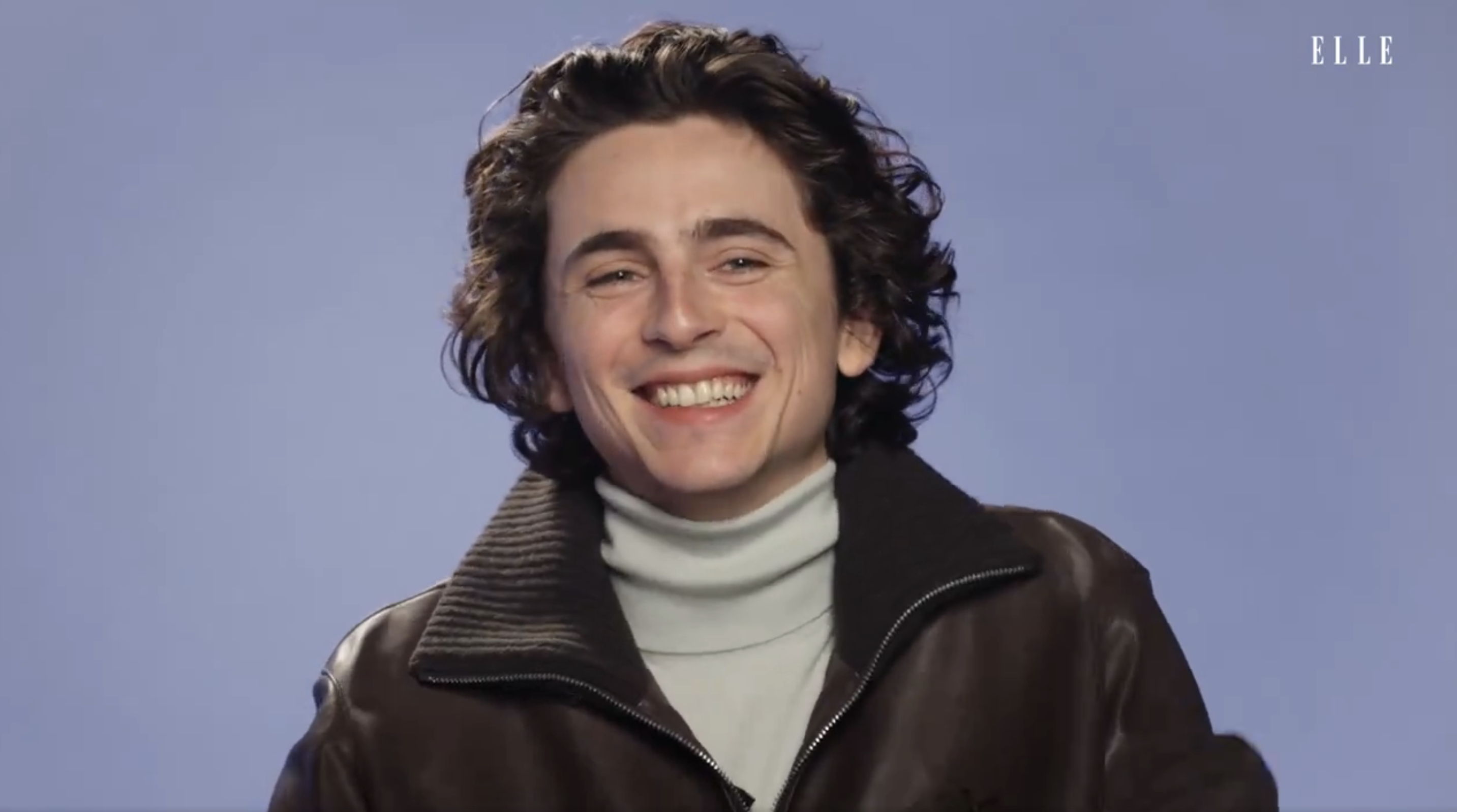 Timothée Chalamet with curly hair wearing a turtleneck and leather jacket smiles at the camera