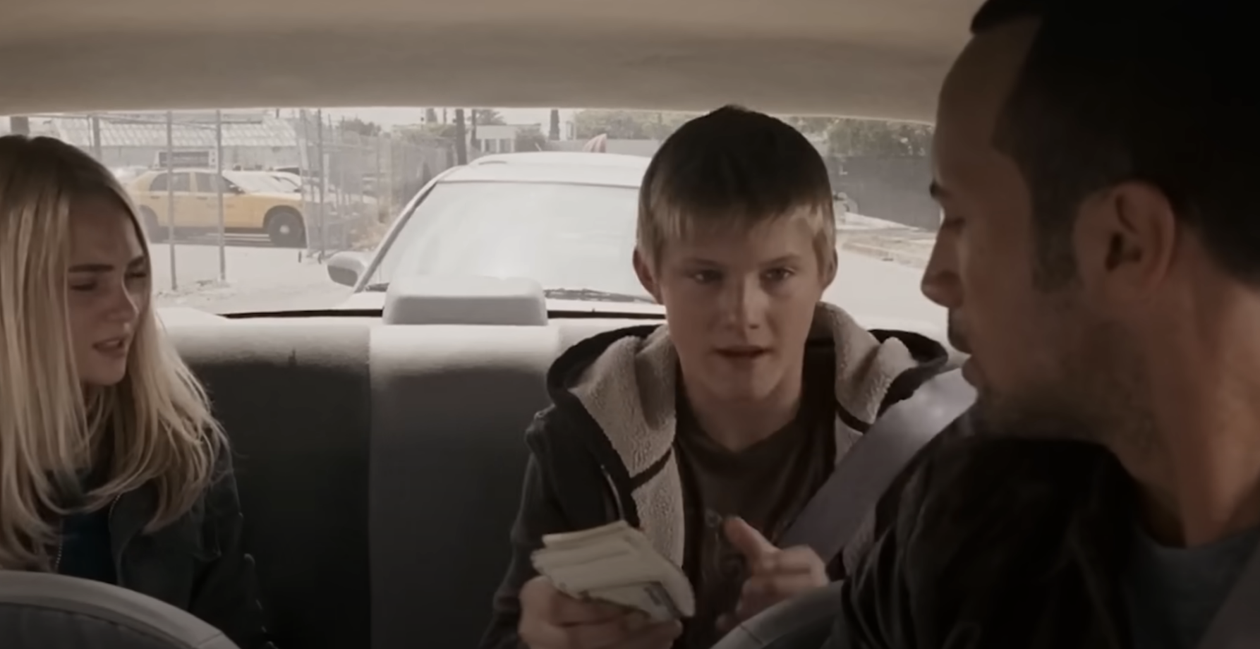 Three characters in a car scene from a movie, with a young boy holding cash in the center