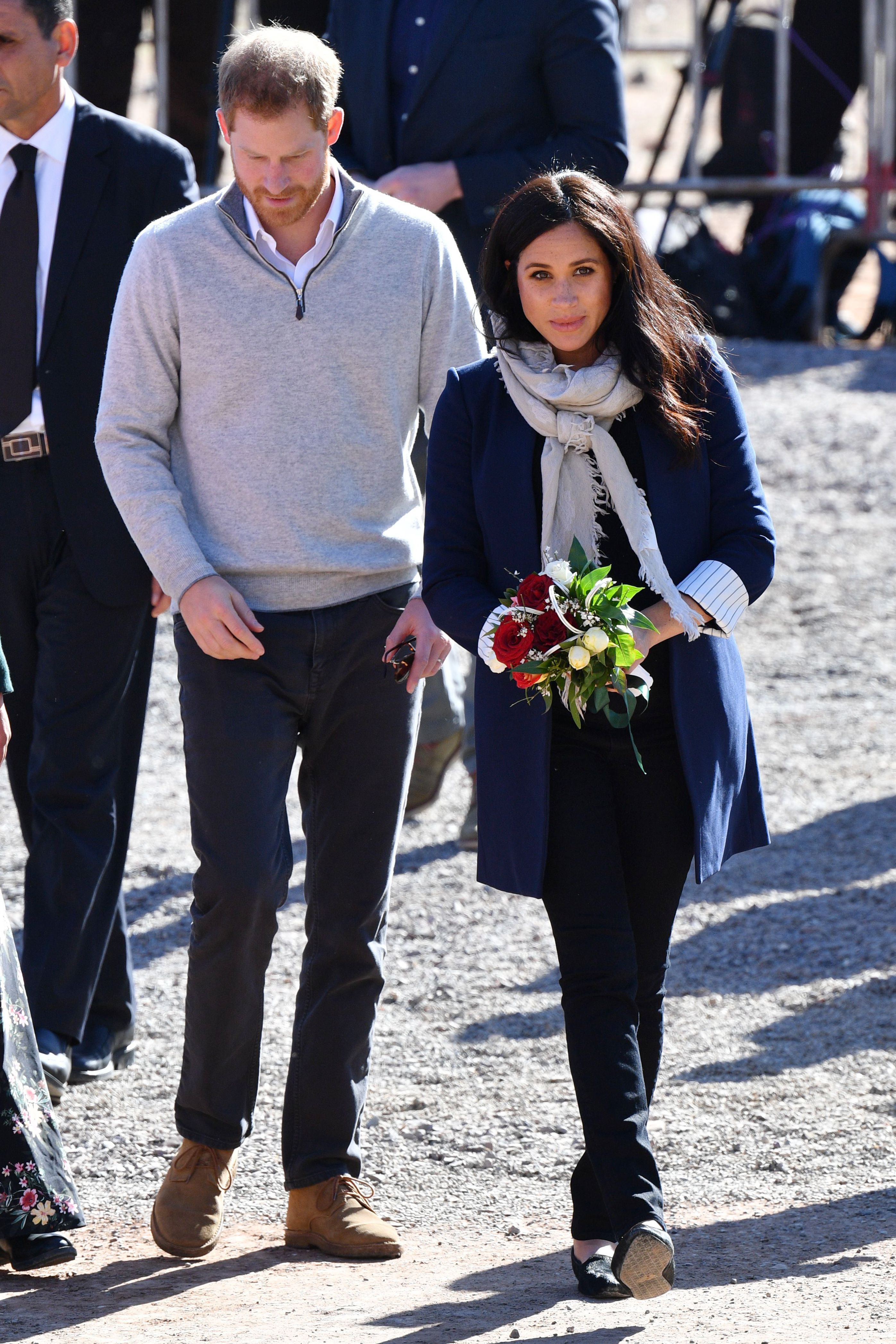 Prince Harry and Meghan Markle walking together, Meghan holding flowers, both dressed in casual attire