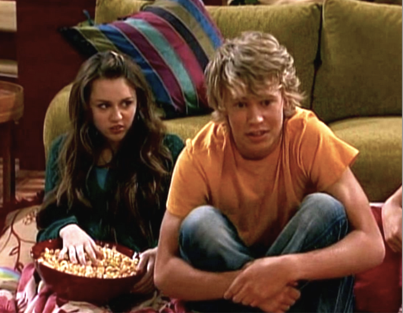 Miley Cyrus and Austin Butler sitting on the ground in front of a couch in a scene from the show. Miley is holding a bowl of popcorn
