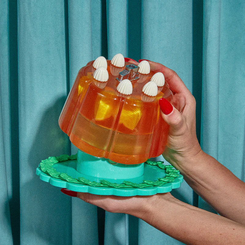 Person holding a whimsical gelatin dessert styled with decorative toppings on a platter, against a draped background