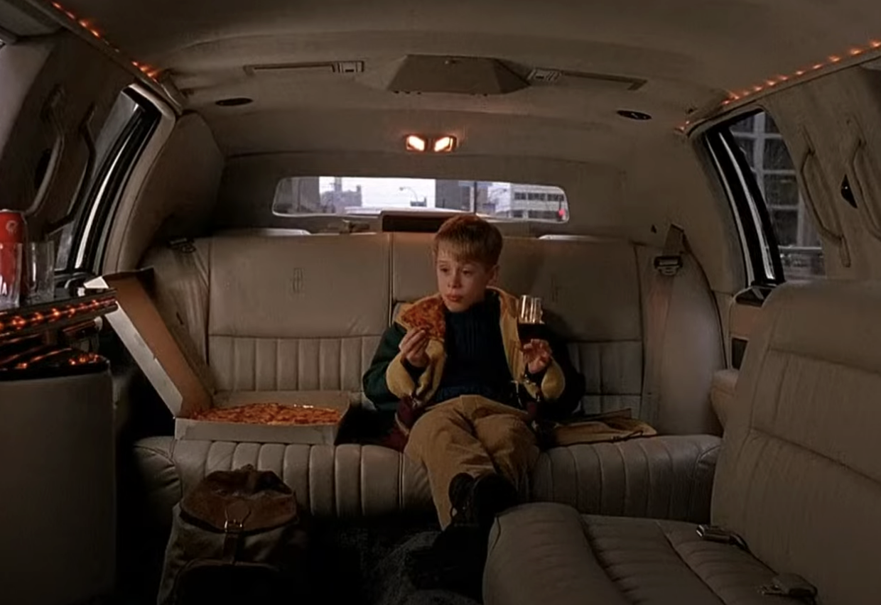 Child character Kevin McCallister sitting in a limousine with a cheese pizza, holding a slice