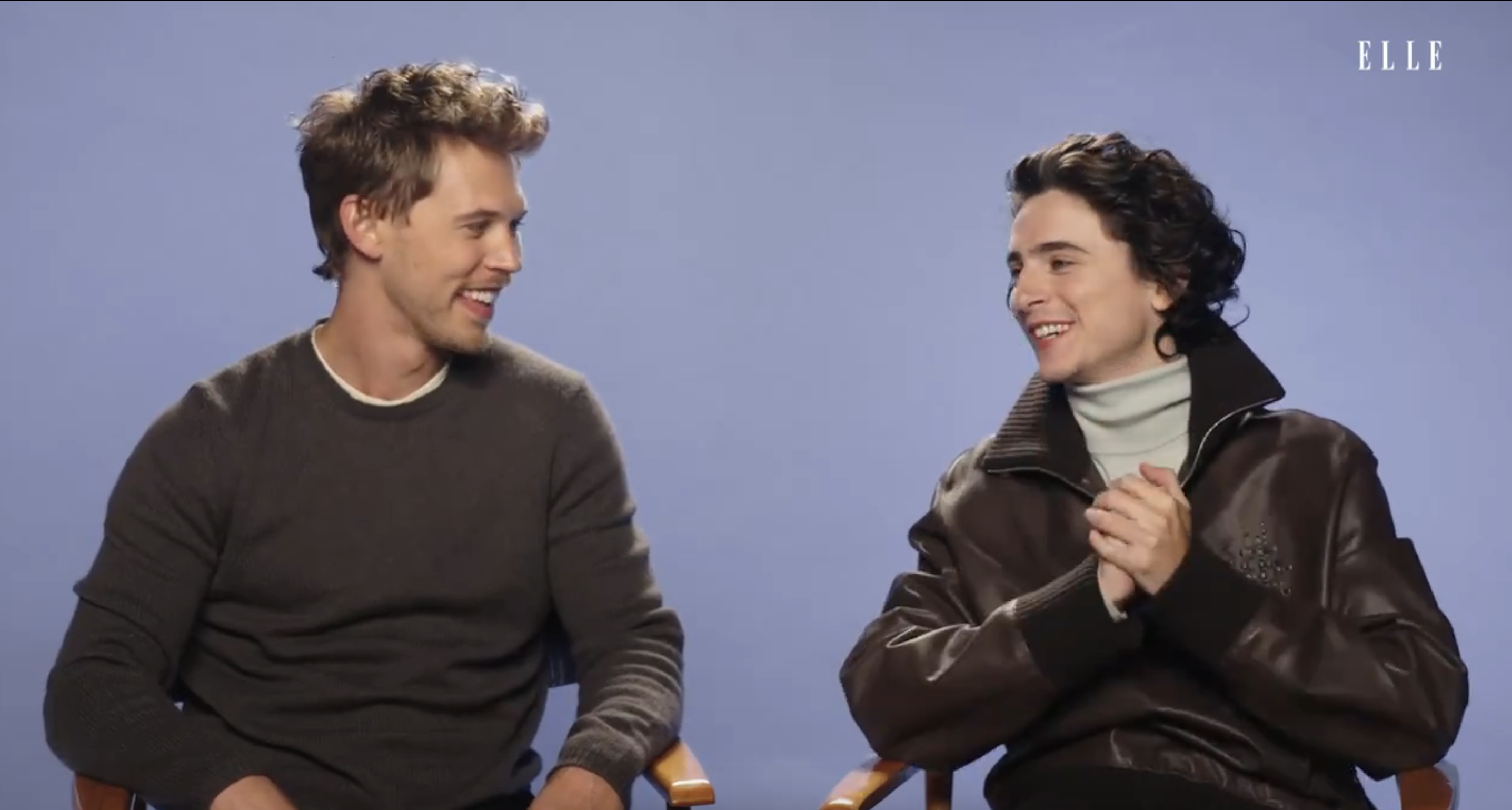Timothée Chalamet and Austin Butler sitting on chairs, engaged in a joyful conversation, with one wearing a turtleneck and leather jacket
