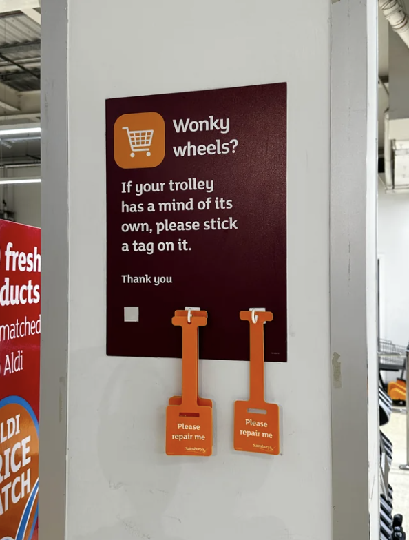 Sign reads &quot;Wonky wheels? If your trolley has a mind of its own, please stick a tag on it&quot; with orange tags labeled &quot;Please repair me&quot; below