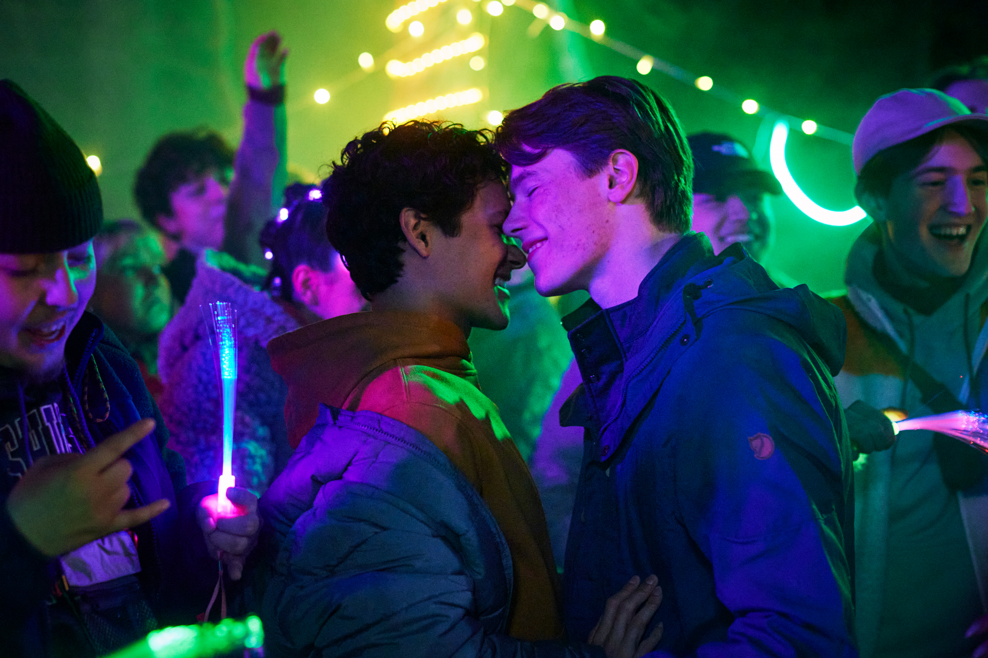 Two actors in a close moment on a TV show set with extras and neon lights in the background