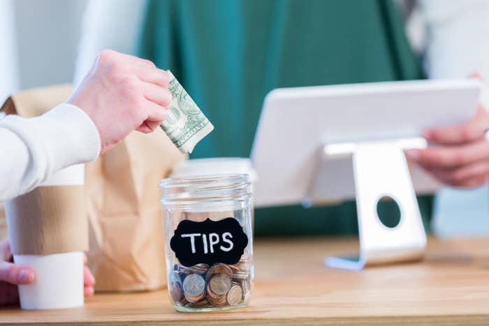 Person putting cash into a tip jar labeled &quot;TIPS&quot; on a counter, beside another person and a POS system