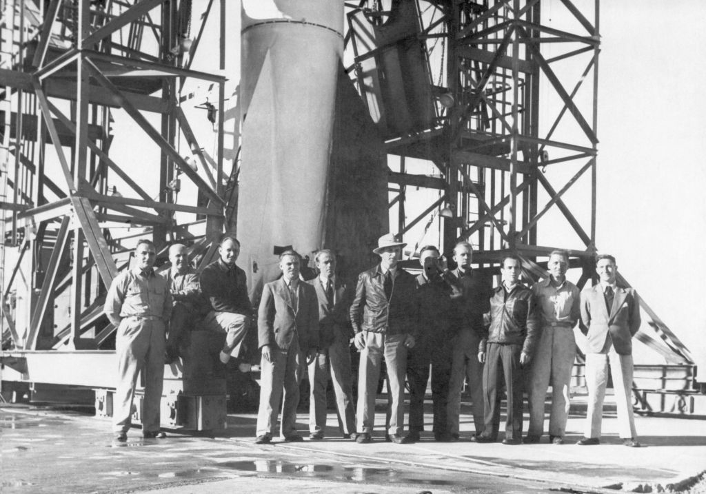 Group of individuals in vintage attire standing in front of a rocket