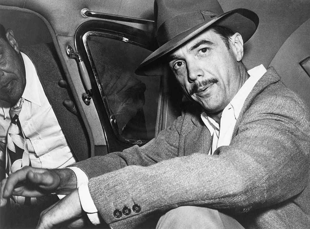 Howard Hughes in a fedora and suit sitting in a car, looking at the camera