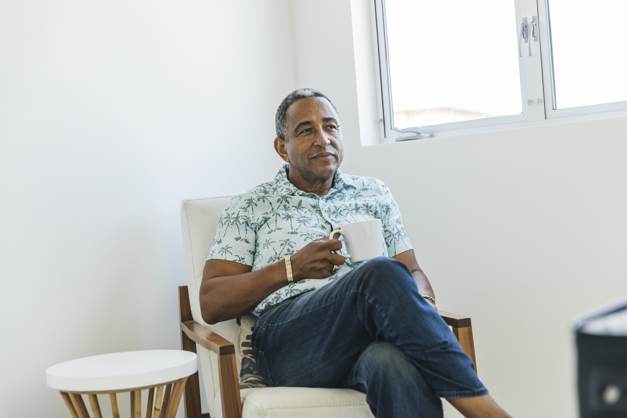 Man sitting in chair by window holding a mug, relaxed posture, in casual attire