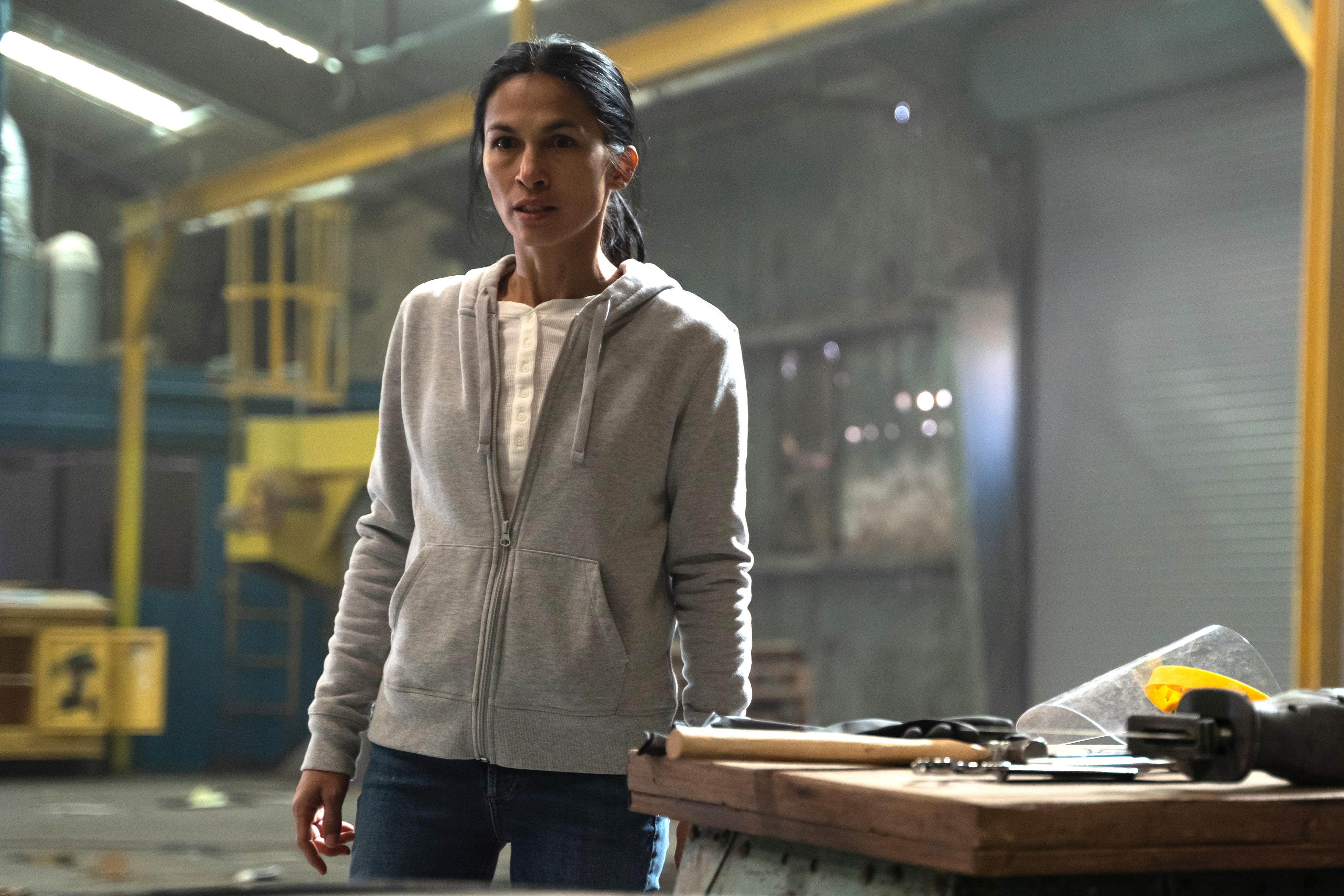 Woman in a casual hoodie stands in an industrial setting, possibly a scene from a TV show or movie