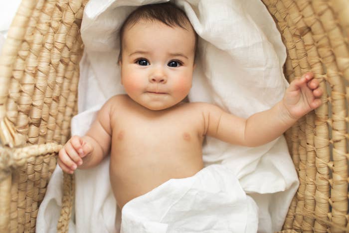 Baby lying in a basket under a light blanket, looking up, one arm extended out