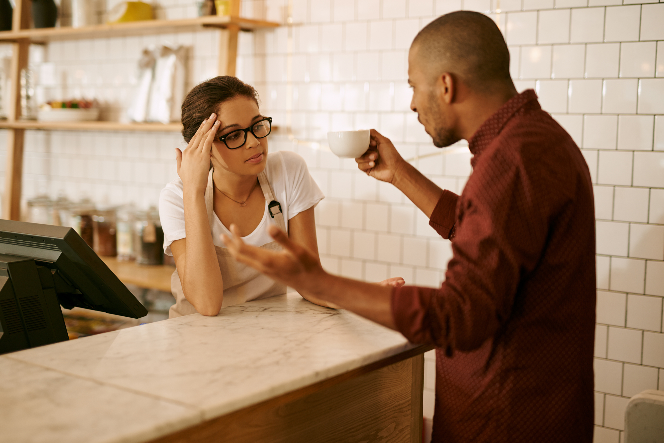 Woman at counter listens to man gesturing with coffee cup, both in casual attire