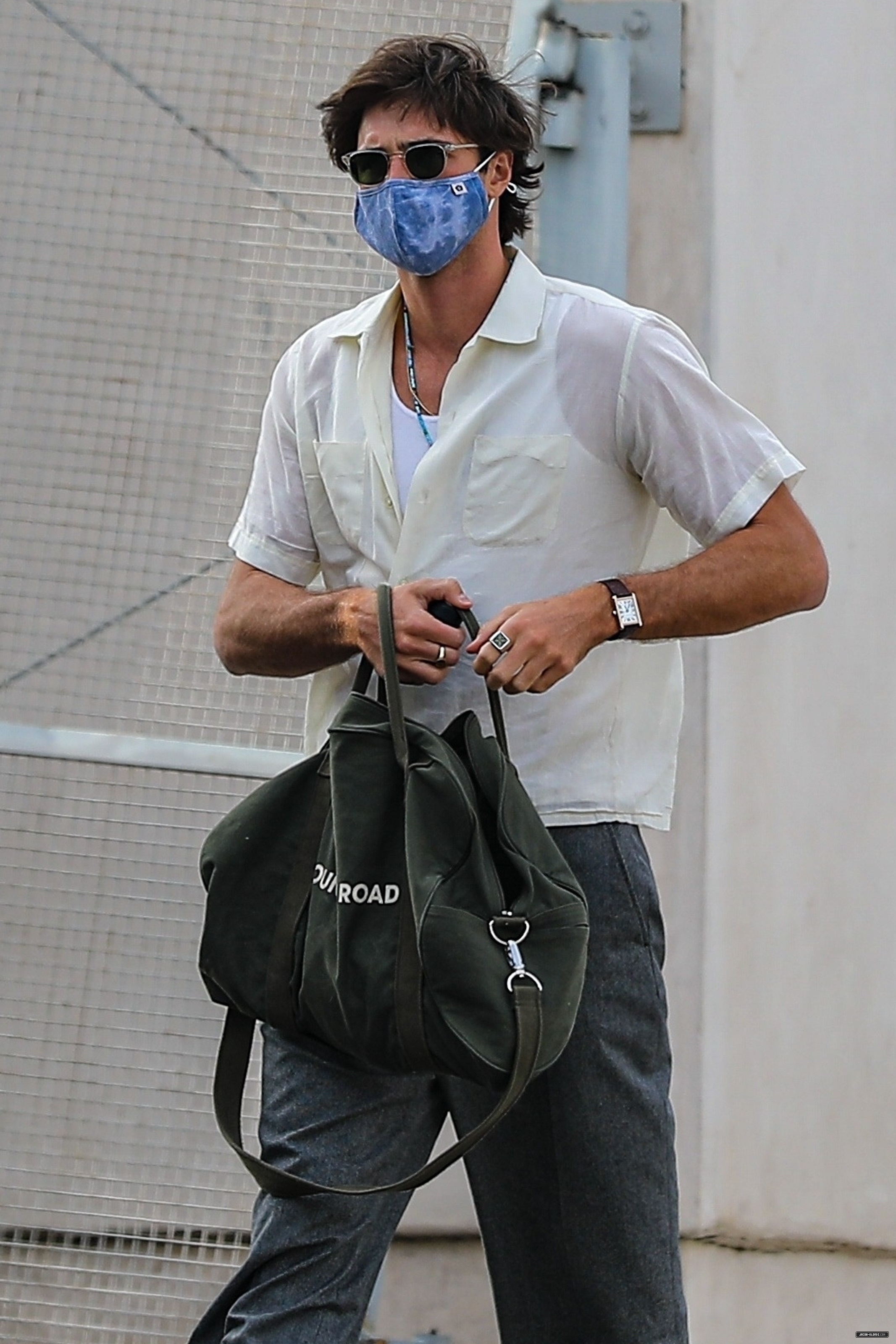 Man in casual shirt and pants with a mask, holding sunglasses and a duffle bag, walking outside
