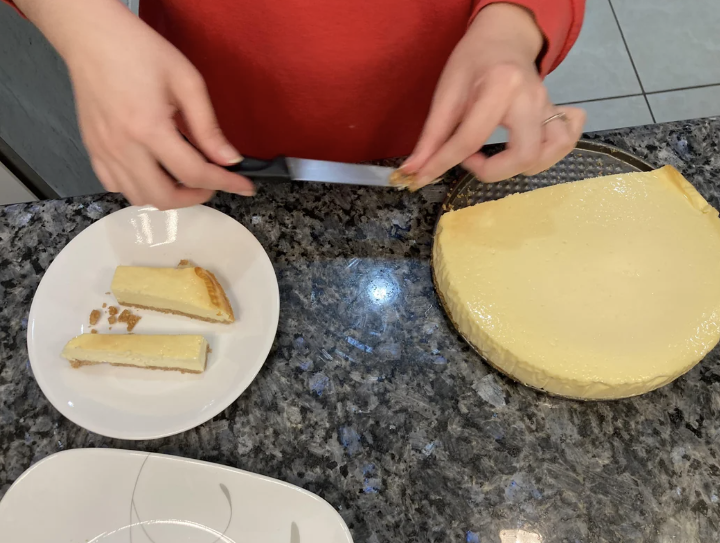 Person slicing a cheesecake badly with two pieces on plates nearby