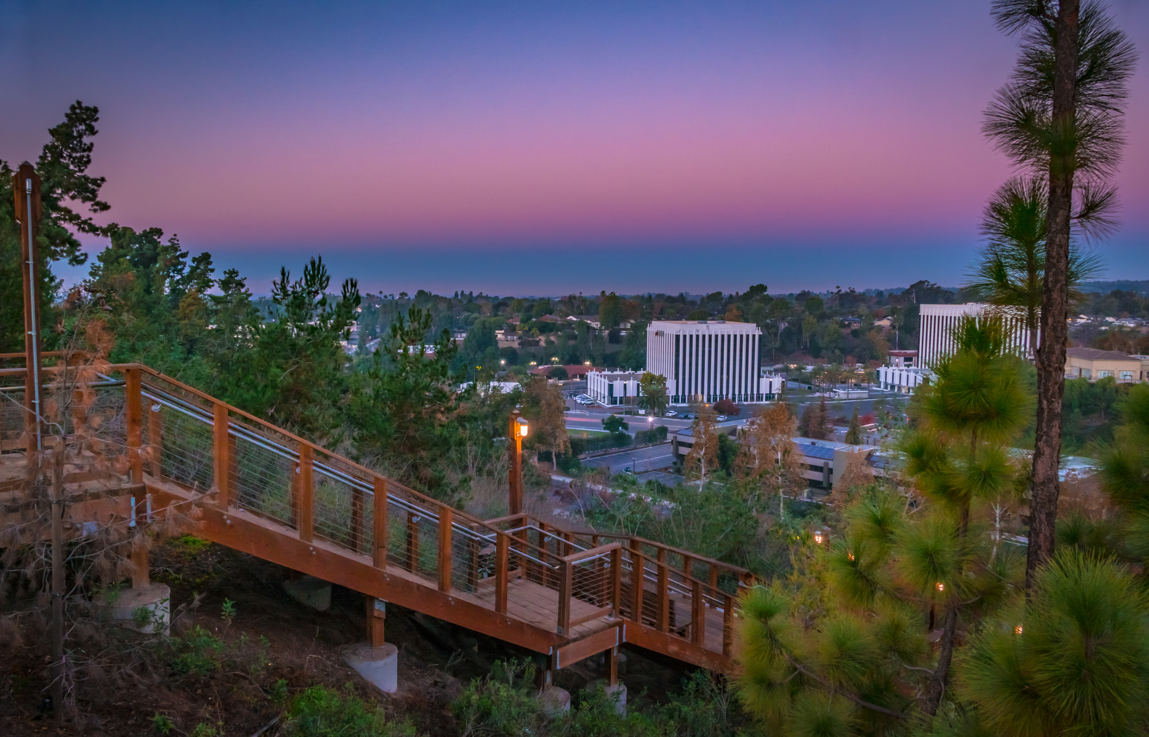 Wooden walkway with handrails leading through a scenic area overlooking a town at twilight