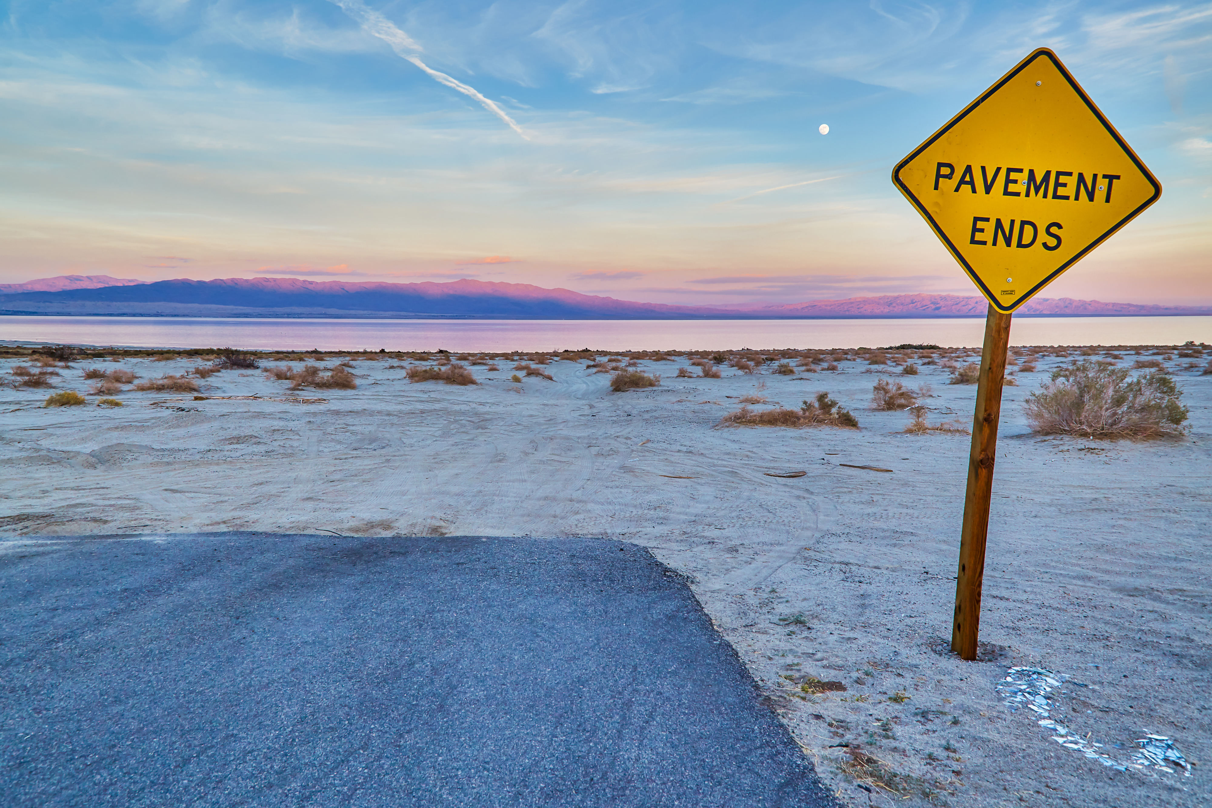 Sign reading &quot;PAVEMENT ENDS&quot; next to a road transitioning into a natural landscape at dusk