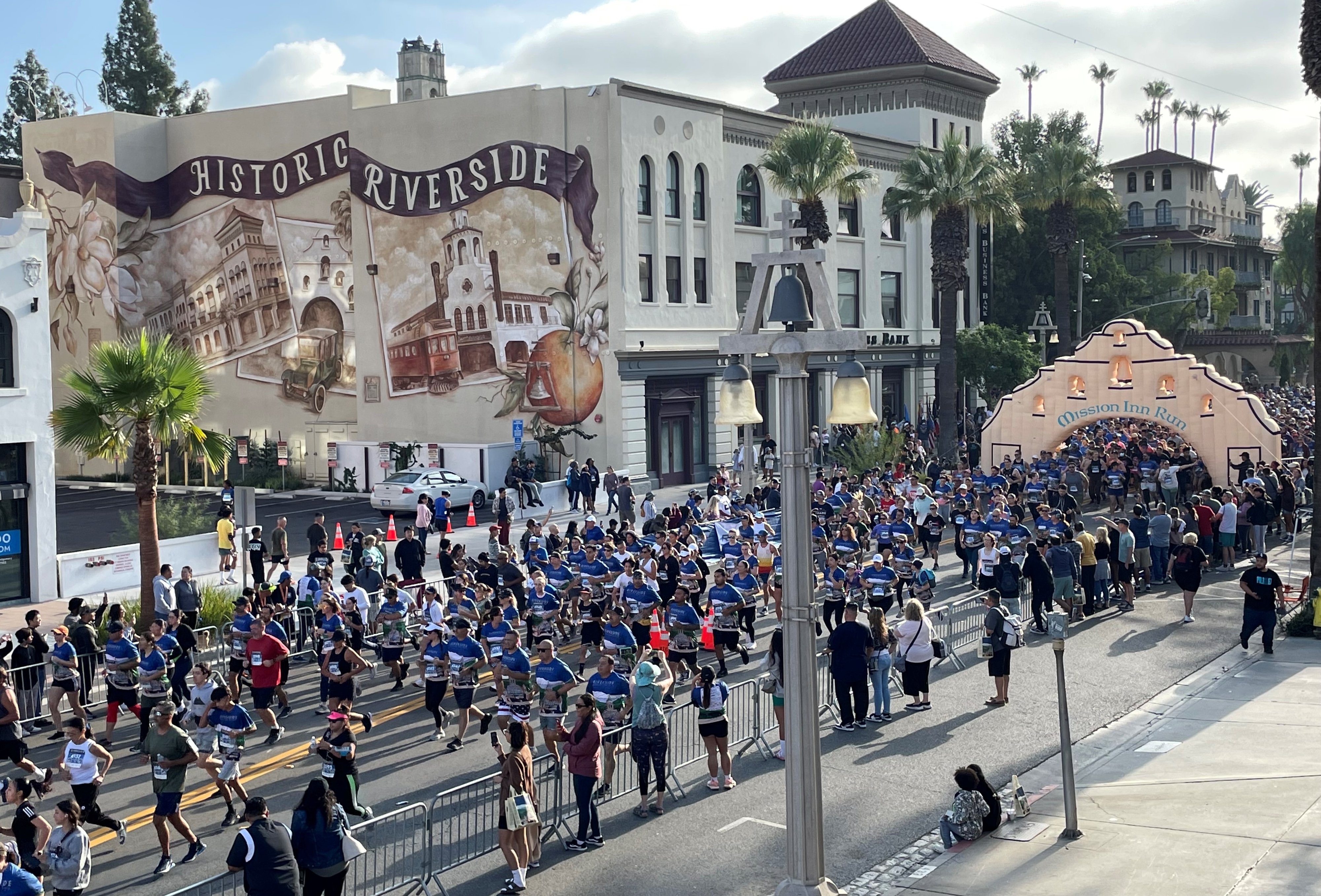 Marching band parades through a city street with onlookers, near buildings with &#x27;Historic Riverside&#x27; mural