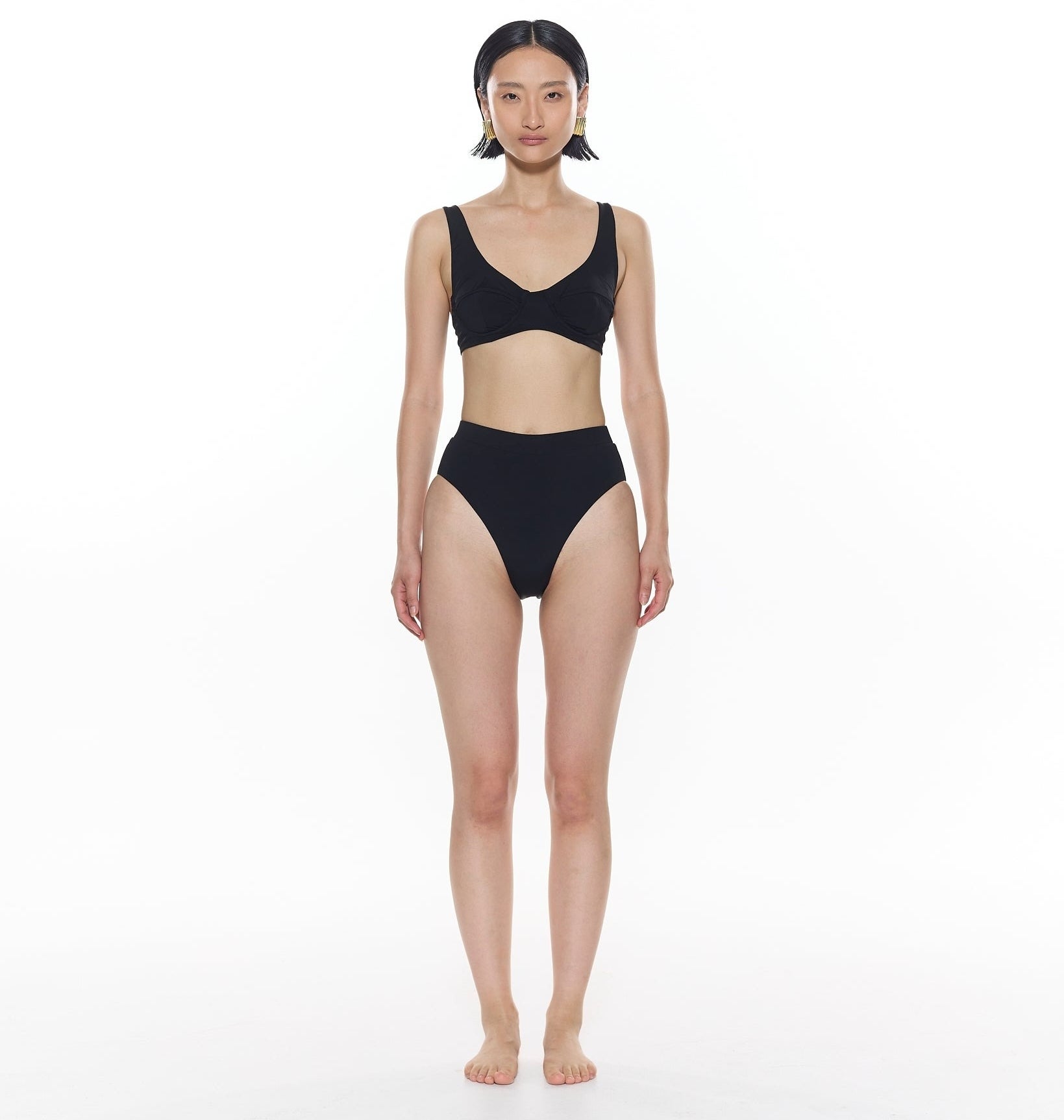 A model standing straight, wearing a simple black two-piece swimsuit, looking directly at the camera