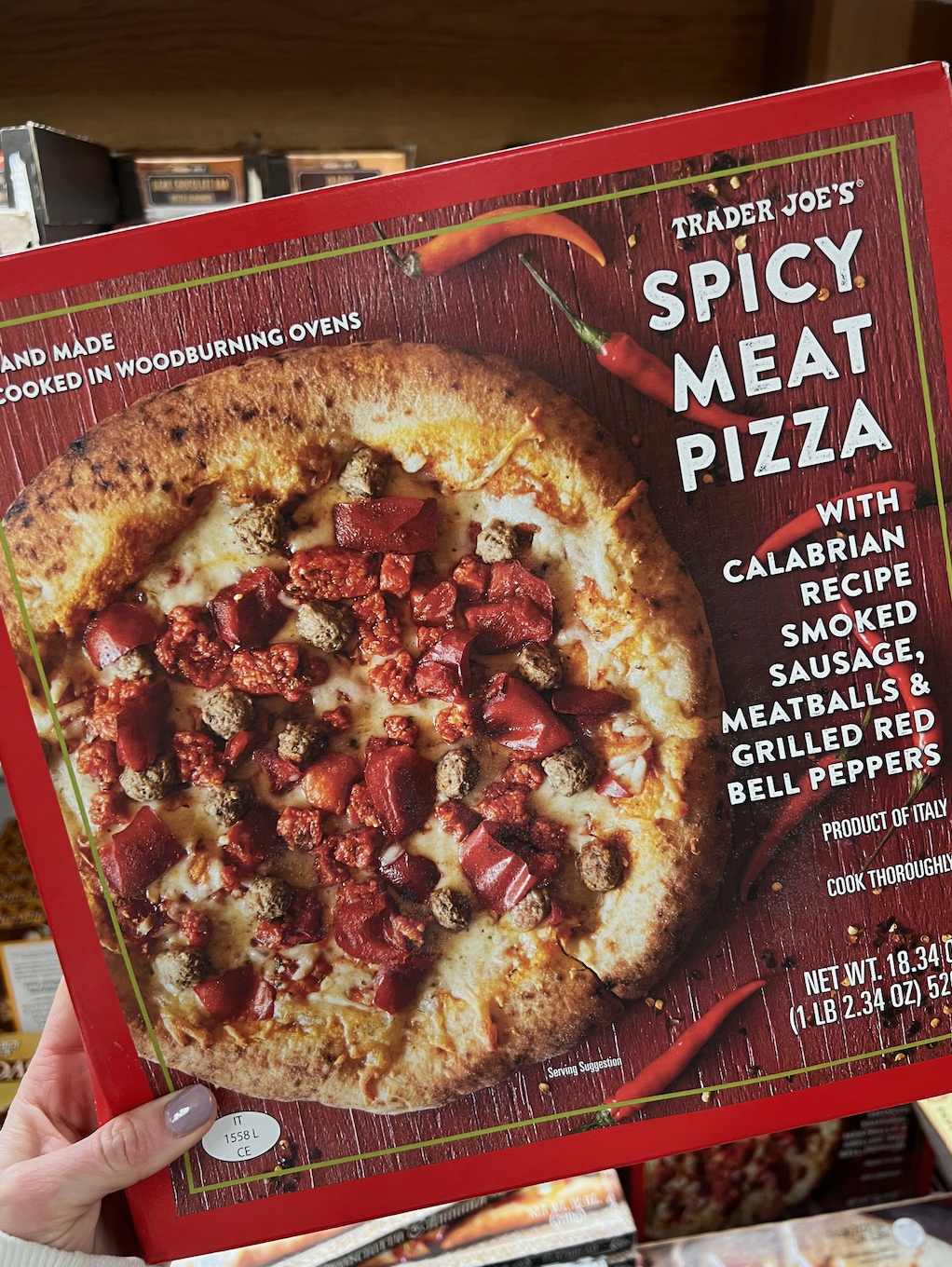 Person holding a Trader Joe&#x27;s Spicy Meat Pizza box showing pizza with Calabrian chili sauce, smoked sausage, meatballs, and red bell peppers
