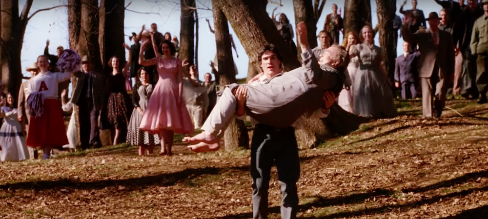 Scene from a film with a man carrying a woman in his arms, others cheering in the background