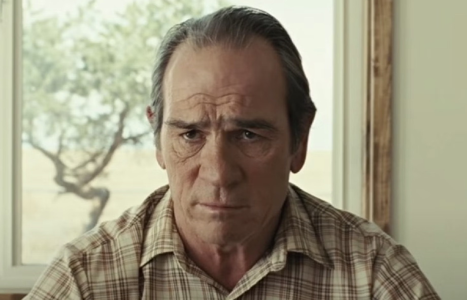Tommy Lee Jones wearing a plaid shirt, seated indoors, with a pensive expression