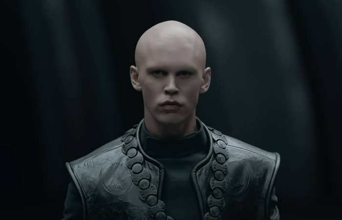 A close-up of Feyd-Rautha with a bald head wearing a detailed high-collared outfit standing with a dark backdrop behind him