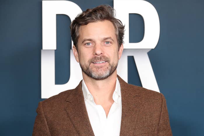 Man in a brown jacket poses before a backdrop with large letters