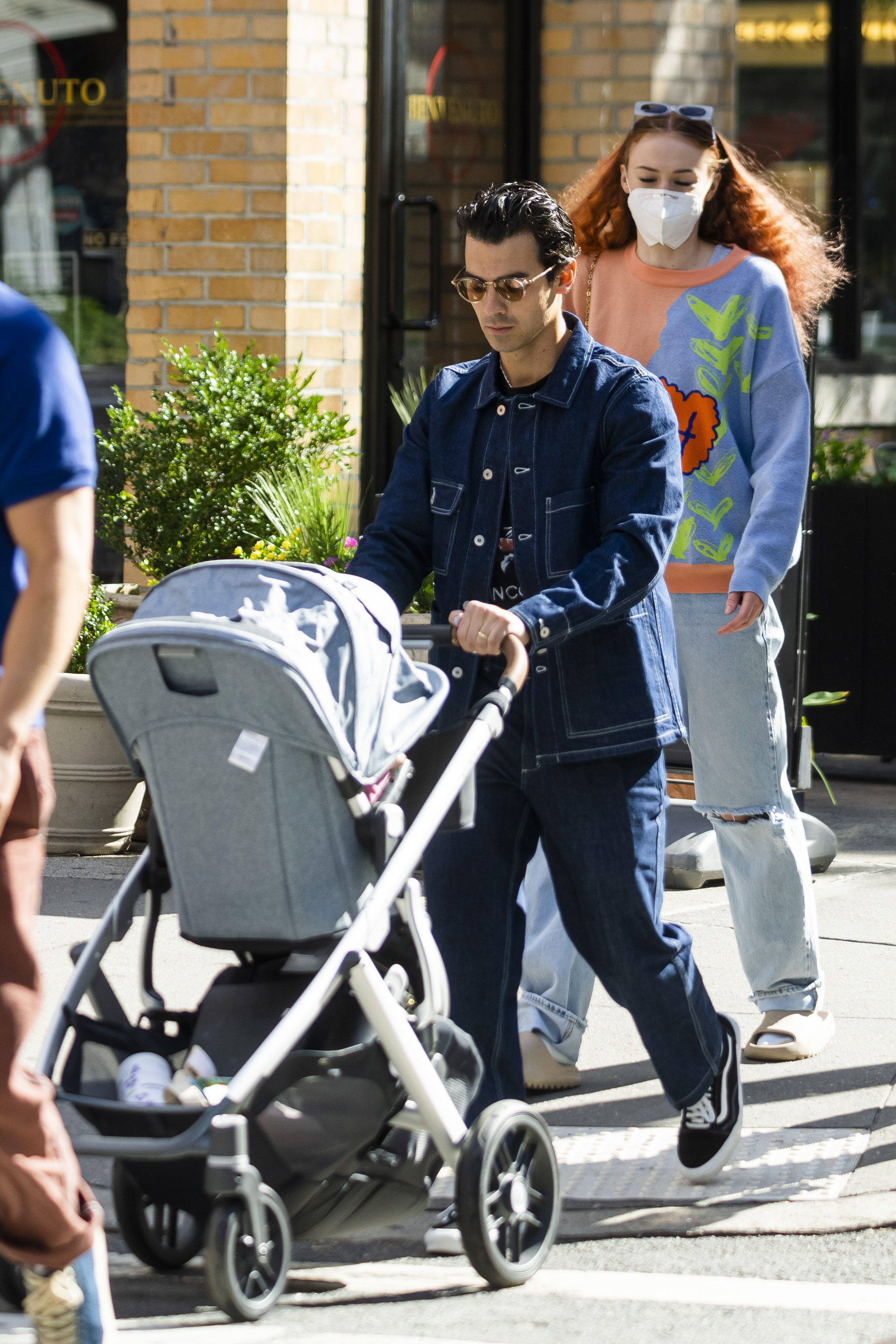 Joe in denim jacket and sunglasses pushes a stroller on city sidewalk as Sophie, who&#x27;s wearing a patterned sweater, follows