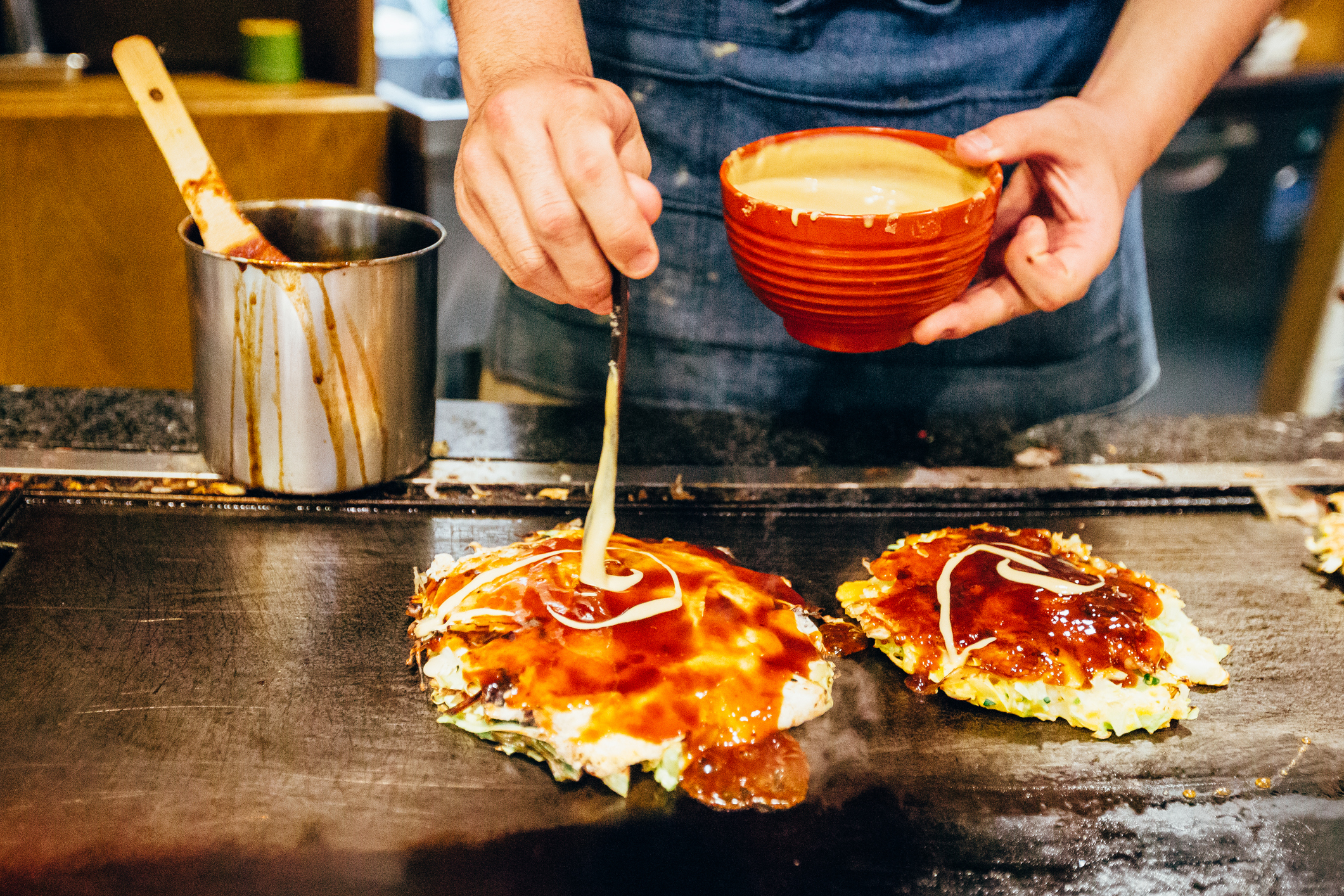 Person in an apron pours sauce on okonomiyaki (Japanese pancakes) at a grill station